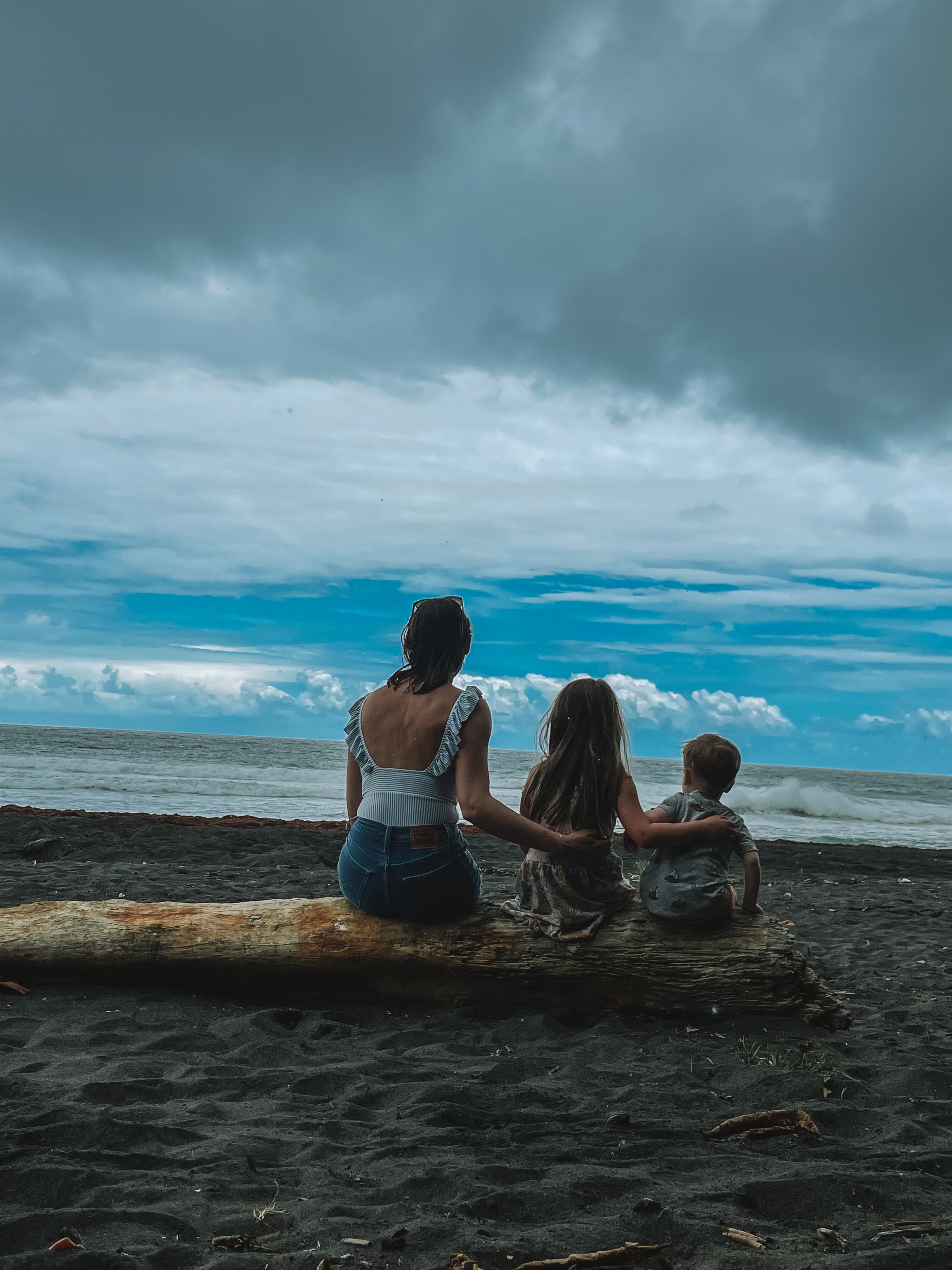 Summer and her two kids sitting on driftwood on a black sand beach in Dominica