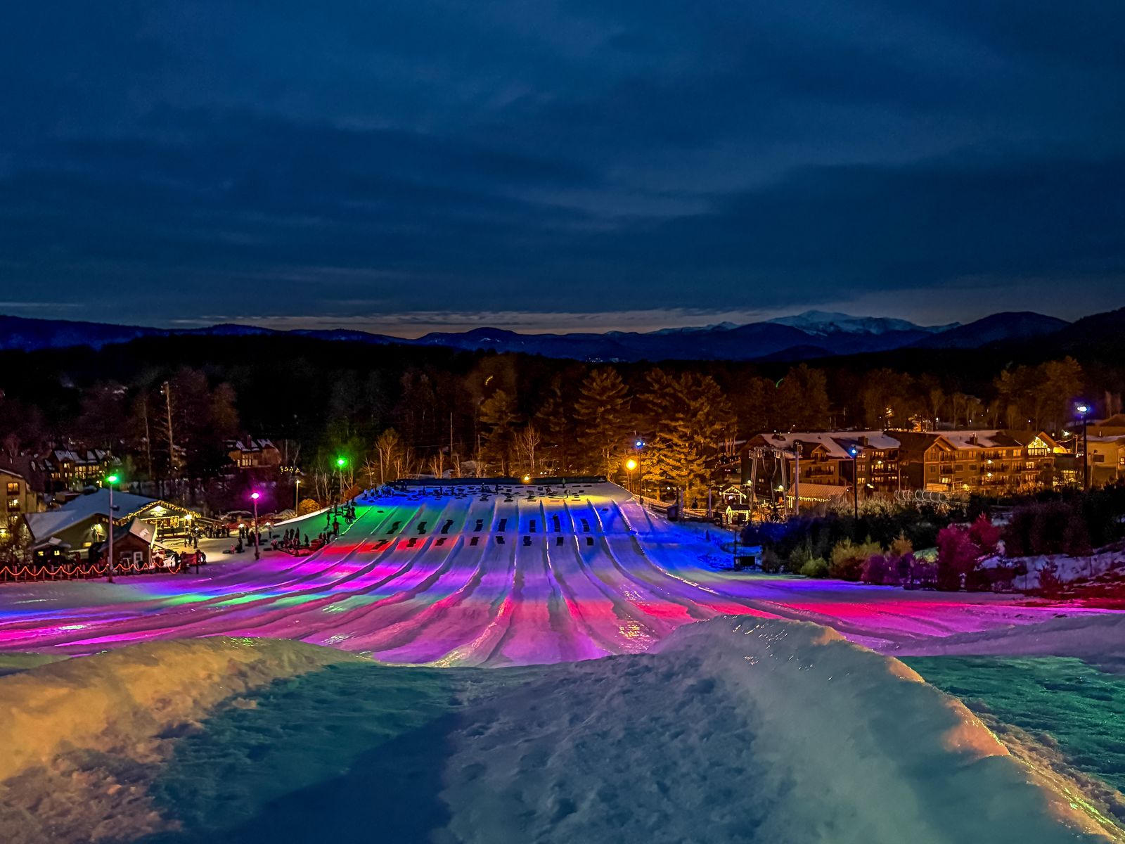 Snow tubing lanes lit pink, blue and purple at night at Cranmore, with North Conway's lights beyond