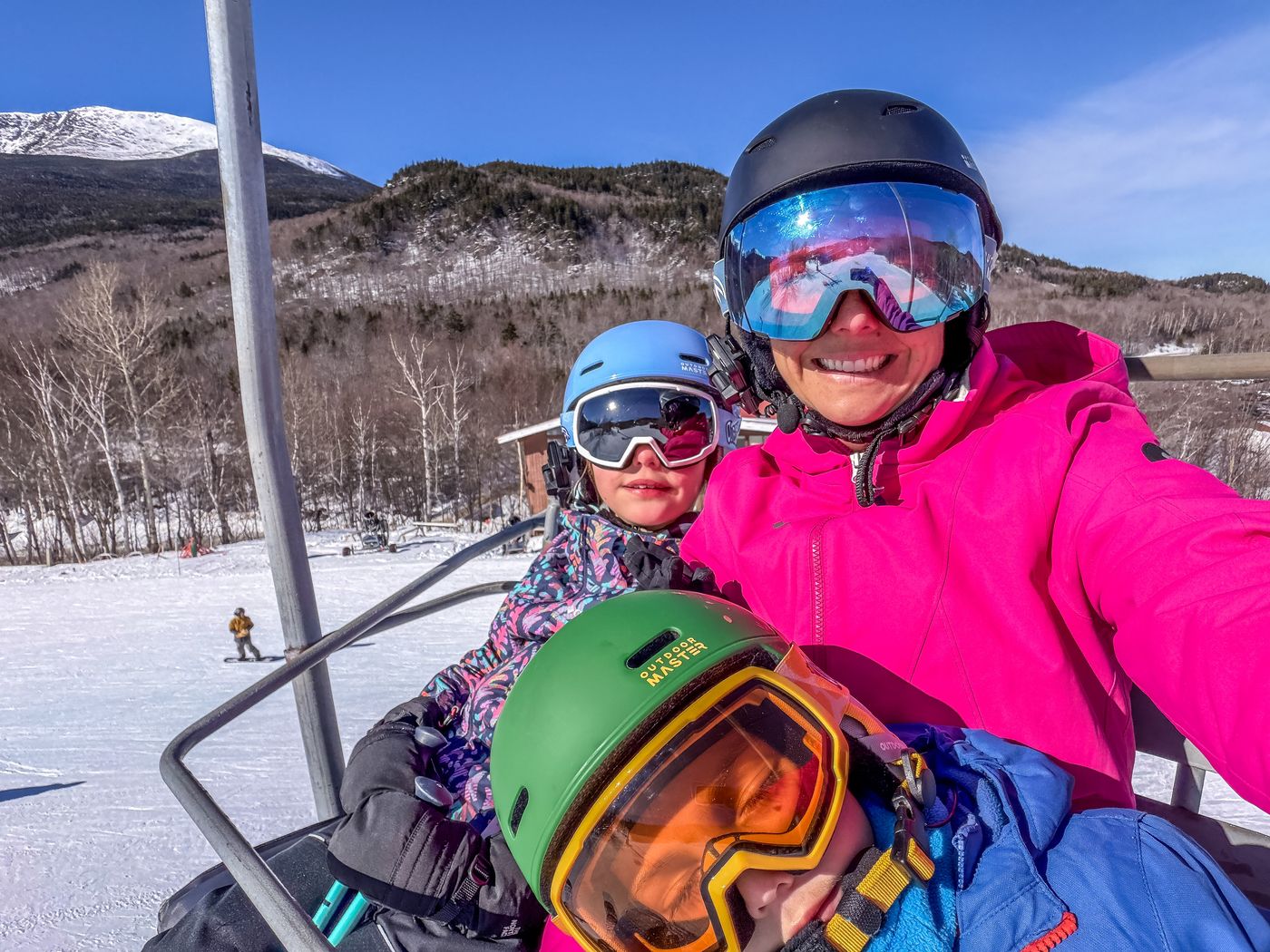 Chairlift selfie of a mom and two kids in helmets and goggles with Mount Washington behind them at Wildcat